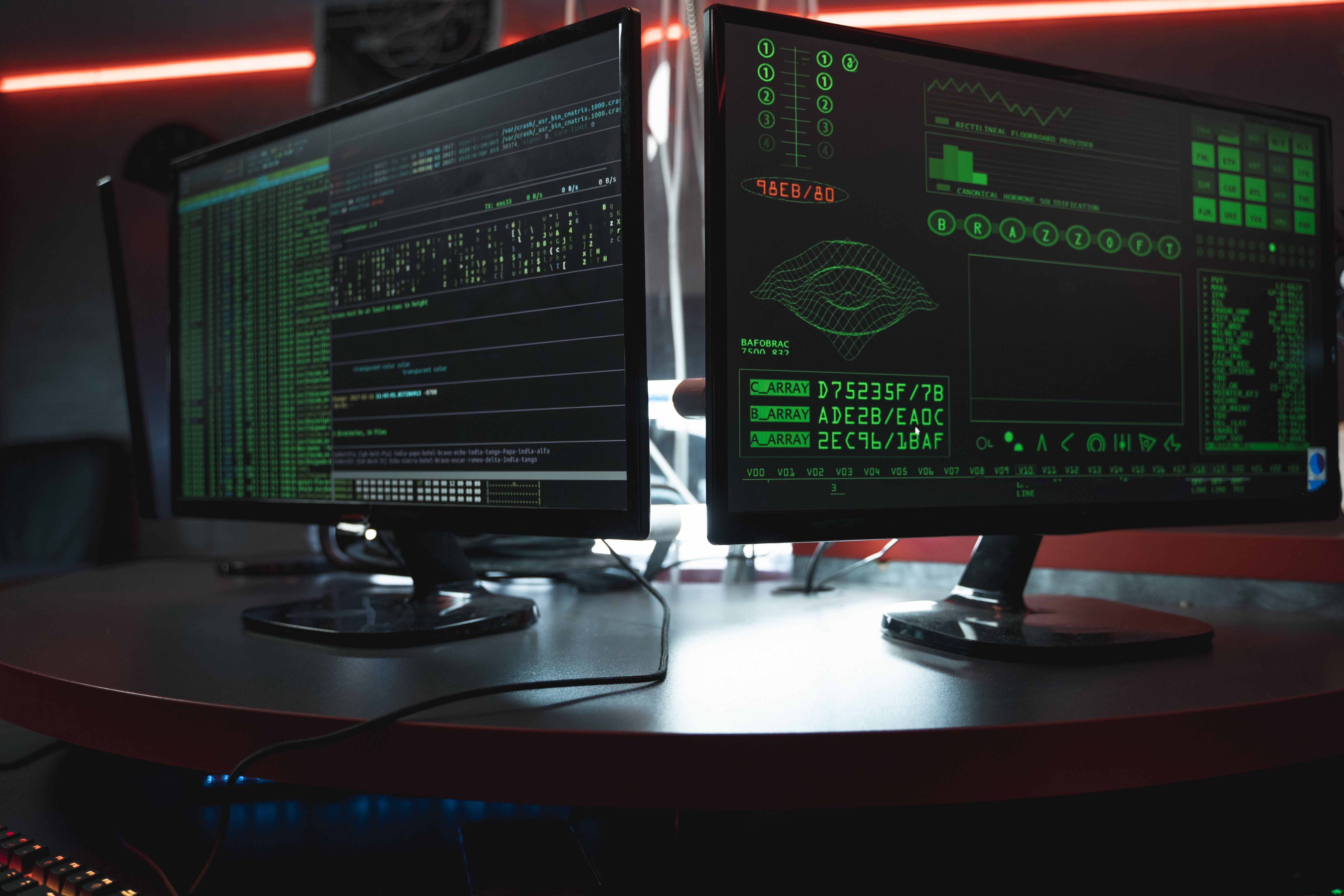 Two computer monitors displaying green code and data visualizations in a dark room.